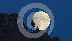 Silhouette of three people on rocks opposite huge moon.