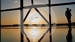 Silhouette of a tourist guy watching the take-off of the plane standing at the airport window at sunset in the evening