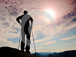 Silhouette of tourist with poles in hand. Hiker stand on rocky view point above misty valley. Sunny daybreak in rocky mountains