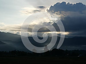 Silhouette of tree and mountain with sunbeam light shoot through the dark cloud to the land at sunrise,