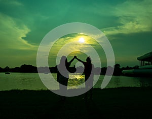 Silhouette of two girls holding hands and raised, glad, in front is a lake, a pond, in a park, in the evening, sunset.