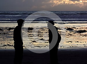 A silhouette of two men enjoying their time and having a conversation on a beach at sunset. The sky is warm orange with some