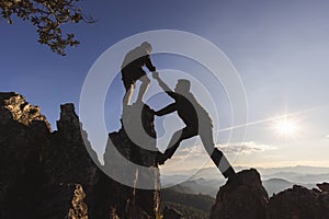Silhouette of  two people climbing on mountain cliff and one of them giving helping hand. People helping and, team work concept