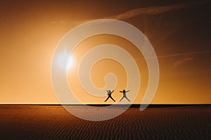 Silhouette of two women friends jumping at the beach at sunset during golden hour. Fun and friendship outdoors