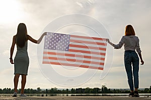 Silhouette of two young friends women holding USA national flag in their hands standing together outdoors. Patriotic girls