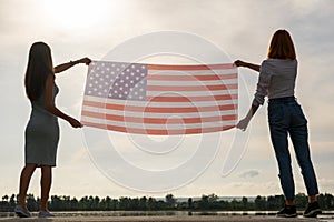 Silhouette of two young friends women holding USA national flag up in their hands standing together. Patriotic girls celebrating