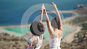 Silhouette of two young girls standing on cliff with the view on blue sea lagoon holding their hands in the air. Women