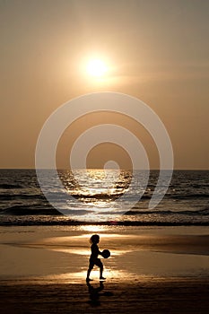 Silhouette of an unrecognizable small child holding a footaball on a golden sandy beach at sunset