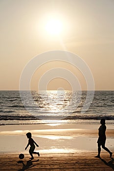 Silhouette of an unrecognizable small child kicking a footaball followed by his mother on a golden sandy beach at sunset