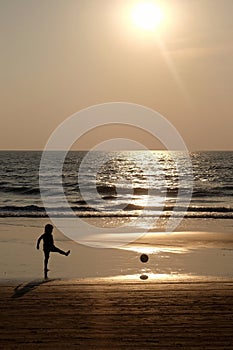 Silhouette of an unrecognizable small child kicking a footaball on a golden sandy beach at sunset