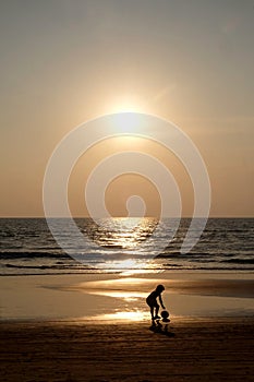 Silhouette of an unrecognizable small child picking up a footaball on a golden sandy beach at sunset