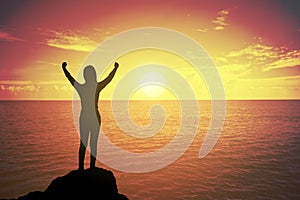 Silhouette of winning success woman at sunset or sunrise standing and raising up hand in celebration of having reached mountain.