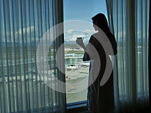 SILHOUETTE: Woman in bathrobe moves the curtain and observes the airport below.