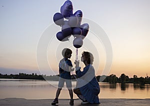 Silhouette of a woman and a child holding a bunch of balloons in their hands, stand by the lake at dusk at sunset