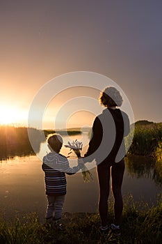 silhouette of woman and child holding each other\'s hands stand with backs near lake against backdrop of fiery sunset