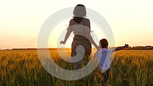 Silhouette of a woman and a child at sunset. A happy mother holds her son's hand and they run across the wheat field.