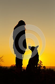 Silhouette woman with a dog looking at each othe rin the field at sunset, pet sitting near girl`s leg on nature,concept of