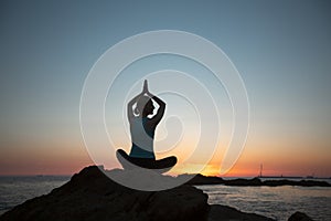 A silhouette of a woman doing yoga, sitting on the shore of the ocean at sunset.