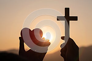 Silhouette of woman hands praying with cross and holding a red heart ball in nature sunrise background, Crucifix, Symbol of