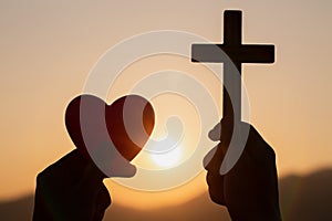 Silhouette of woman hands praying with cross and holding a red heart ball in nature sunrise background, Crucifix, Symbol of