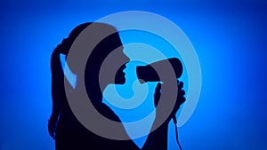Silhouette of woman having fun singing into hair dryer. Female dancing silly on blue background