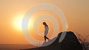 Silhouette of a woman hiker climbing alone down from a big stone at sunset in mountains. Female tourist standing up on high rock i