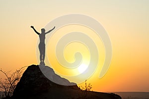 Silhouette of a woman hiker standing alone on big stone at sunset in mountains. Female tourist raising her hands up on high rock