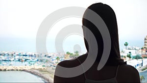 Silhouette of woman open curtains and enjoy sea view from terrace. Female traveler look at marina