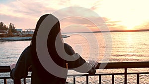 Silhouette of a woman on the pier against the sun. A pensive young girl stands on the beach at the sunset time looks straight and
