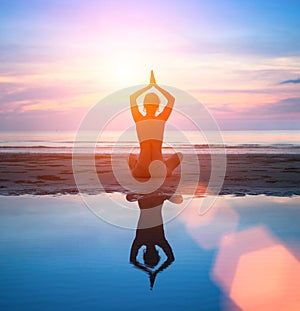 Silhouette of a woman practicing yoga on the beach with reflection in water.