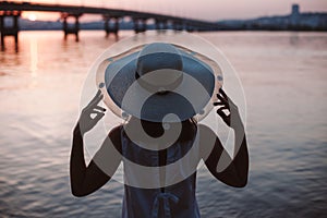 The silhouette of a woman by the river at sunset. Rear view of the silhouette of a woman in a straw hat enjoying the