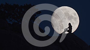 Silhouette of woman siting on rocks opposite huge moon. Lady looking