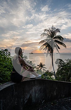 Silhouette of woman sitting alone on the hill with a coconut palm trees and enjoys sea on a horizon in a sunset