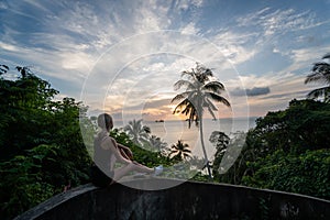 Silhouette of woman sitting alone on the hill with a coconut palm trees and enjoys sea on a horizon in a sunset