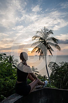 Silhouette of woman sitting alone on the hill with a coconut palm trees and enjoys sea on a horizon in a sunset