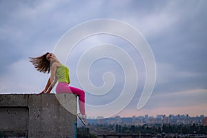 Silhouette of woman sitting on the edge on the dramatic cloudscape during sunset with cityscape on background. concept