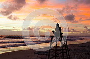 A silhouette woman sitting on a lifeguard tower enjoying the sunrise on a beach and the sun`s rays painting the sky