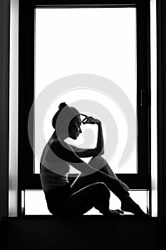 Silhouette of a woman sitting thinking on a window sill. Black and white photography. loneliness