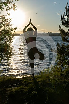 Silhouette of a woman in sportswear standing in the yoga pose Vrikshasana, tree pose on a rug on the lake shore