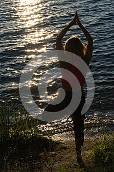 Silhouette of a woman in sportswear standing in the yoga pose Vrikshasana, tree pose on a rug on lake shore. Meditation