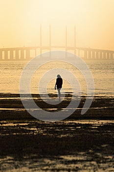 Silhouette of a woman standing on the beach during bright yellow sunset with a bridge in background