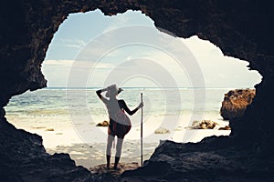 Silhouette of woman standing in cave on the beach with hat, stick and backpack