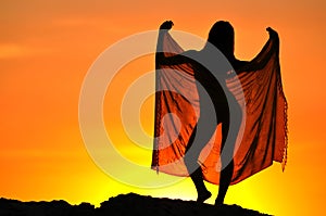 Silhouette of woman standing and holding pareo in raised hands at sunset