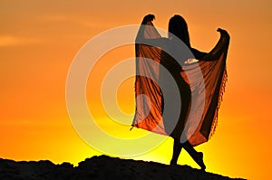 Silhouette of woman standing and holding pareo in raised hands at sunset