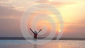 Silhouette of woman in swimsuit and sunglasses, sitting and relaxing with hands up, on edge of outdoor infinity pool