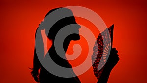 Silhouette of woman use hand fan. Black contur shadow of female`s face in profile on red background