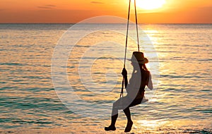 Silhouette woman wear bikini and straw hat swing the swings at the beach on summer vacation at sunset. Enjoying and relaxing girl