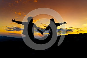 Silhouette of Young Asian Couple Holding Hand at Sunset on Mountain with Orange Sky. Enjoying Peaceful Moment Concept. Relaxing or