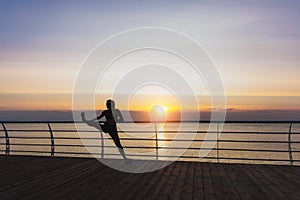 Silhouette of young beautiful athletic girl with long blond hair in headphones listening to music and doing stretching at sunrise
