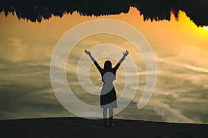 Silhouette of a young beautiful girl with hands up against the background of the sunset in the reflection of the pond